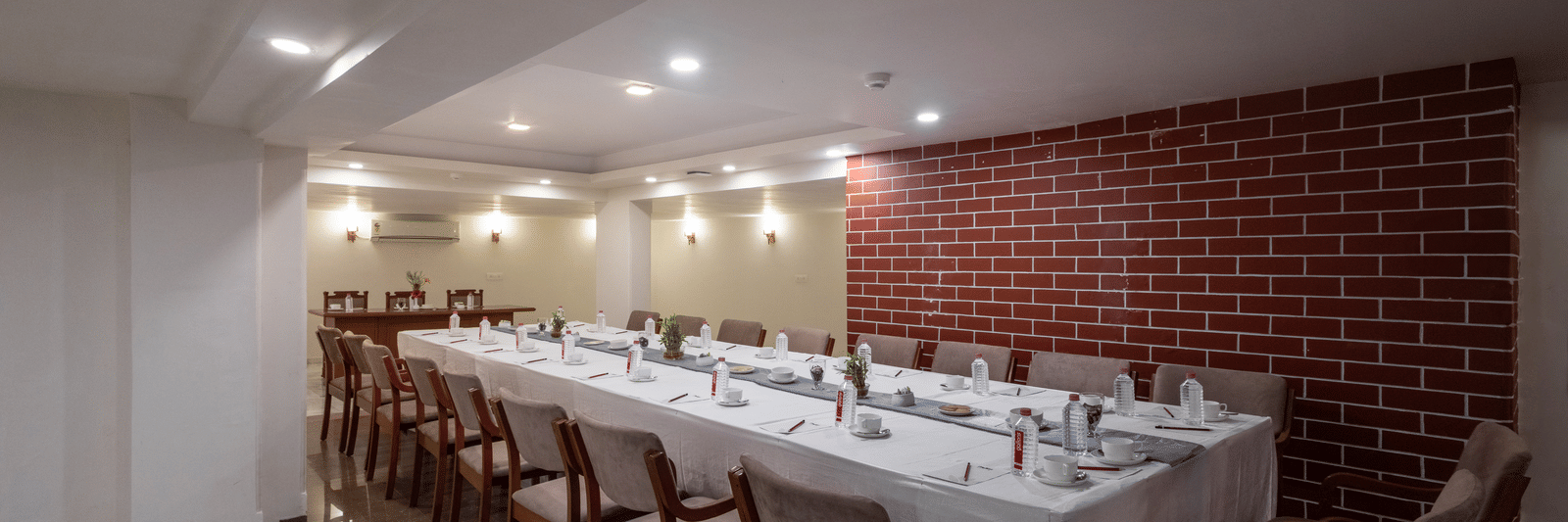 Long, formal dining hall with a table draped in white linen, wooden chairs, and a prominent red brick feature wall at Pravasa Stay, Vadodara.