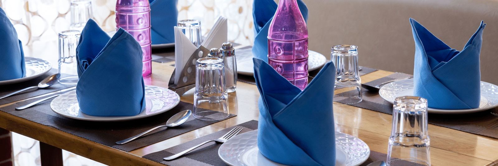Close-up of a wooden table setting with white plates and striking blue and pink folded napkins in a well-lit dining area at Pravasa Stay, Vadodara.