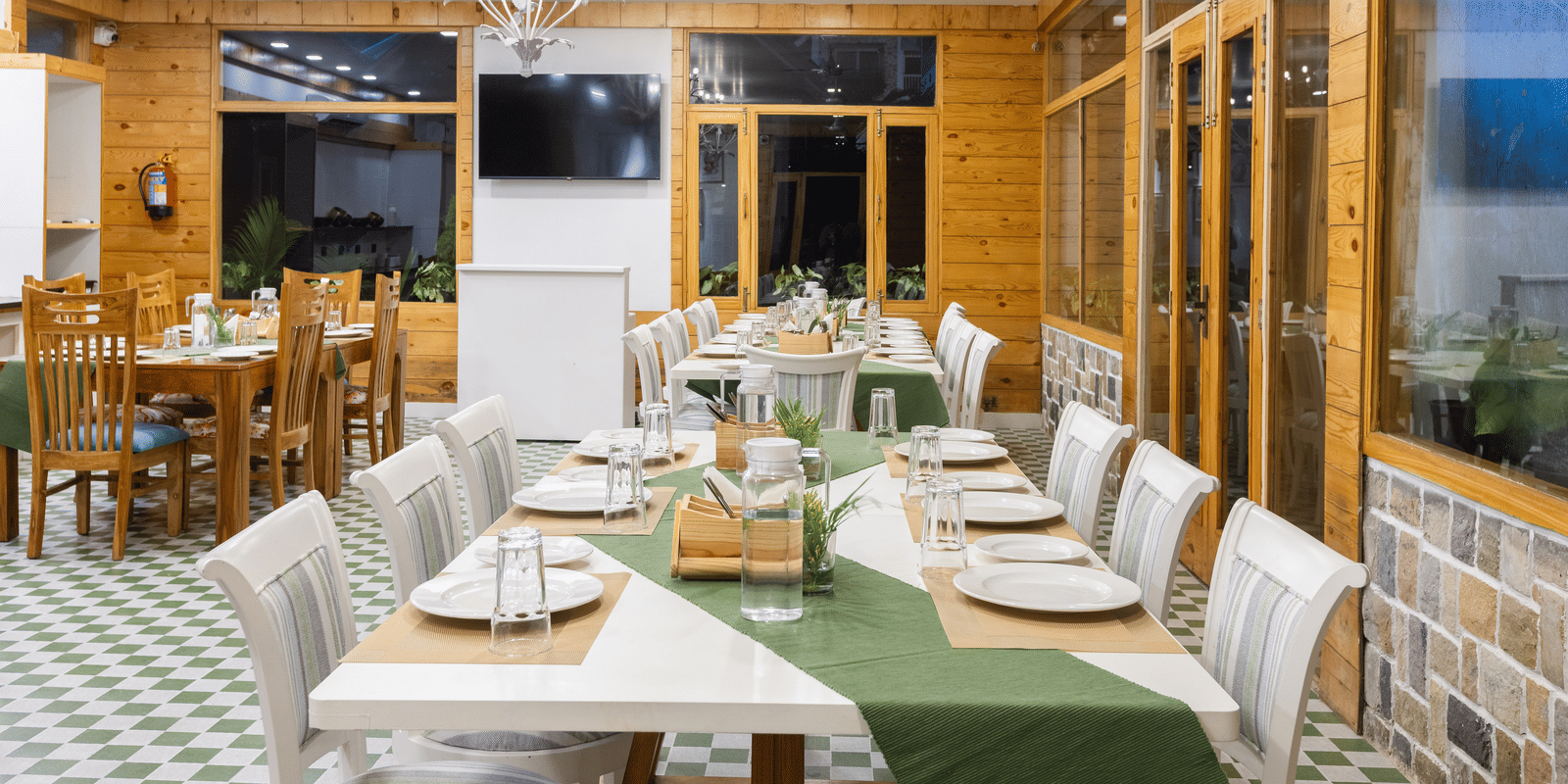 A large dining room with a wooden interior, a long set table, and windows opening to a green view at Megha Resort & Villas, Dehradun.