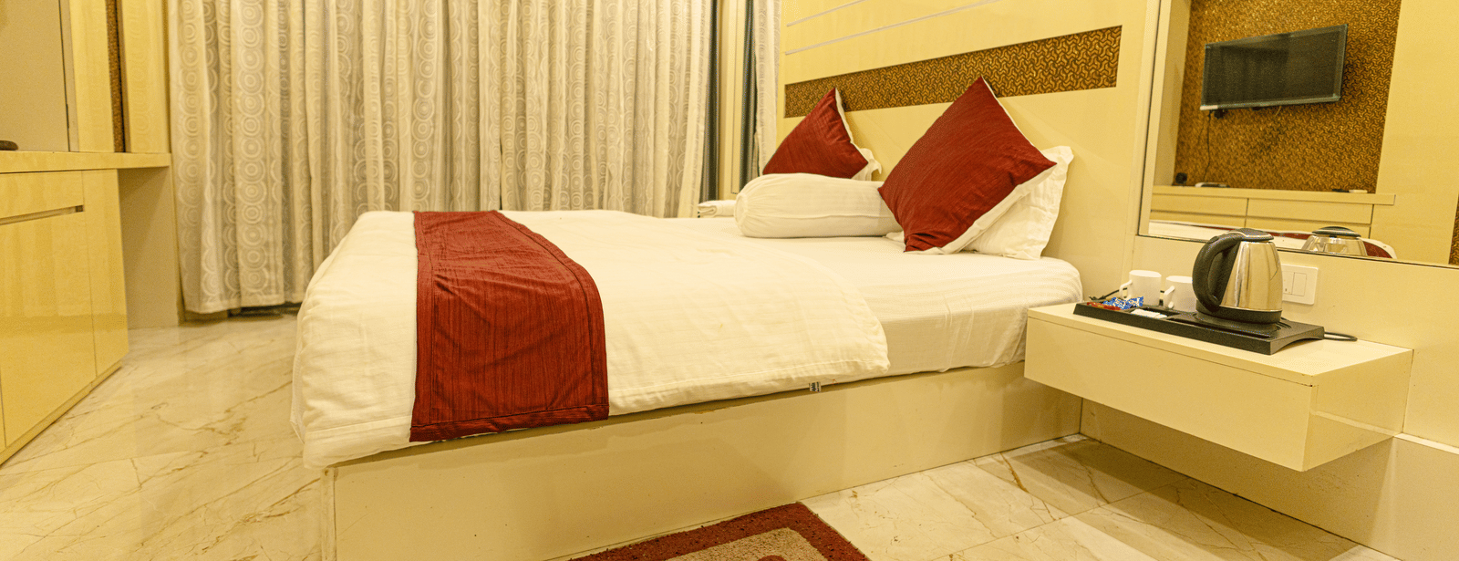 A bedroom with a king size bed, a bedside table with an electric kettle, a television, and curtains at Hotel Seagull Digha.