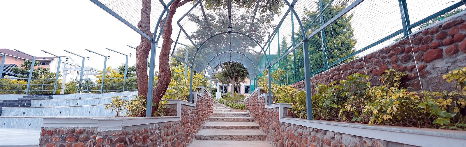 Brick pathway lined with trees and metal archway leading to garden area at Trishvam Symphony, Krishnagiri.