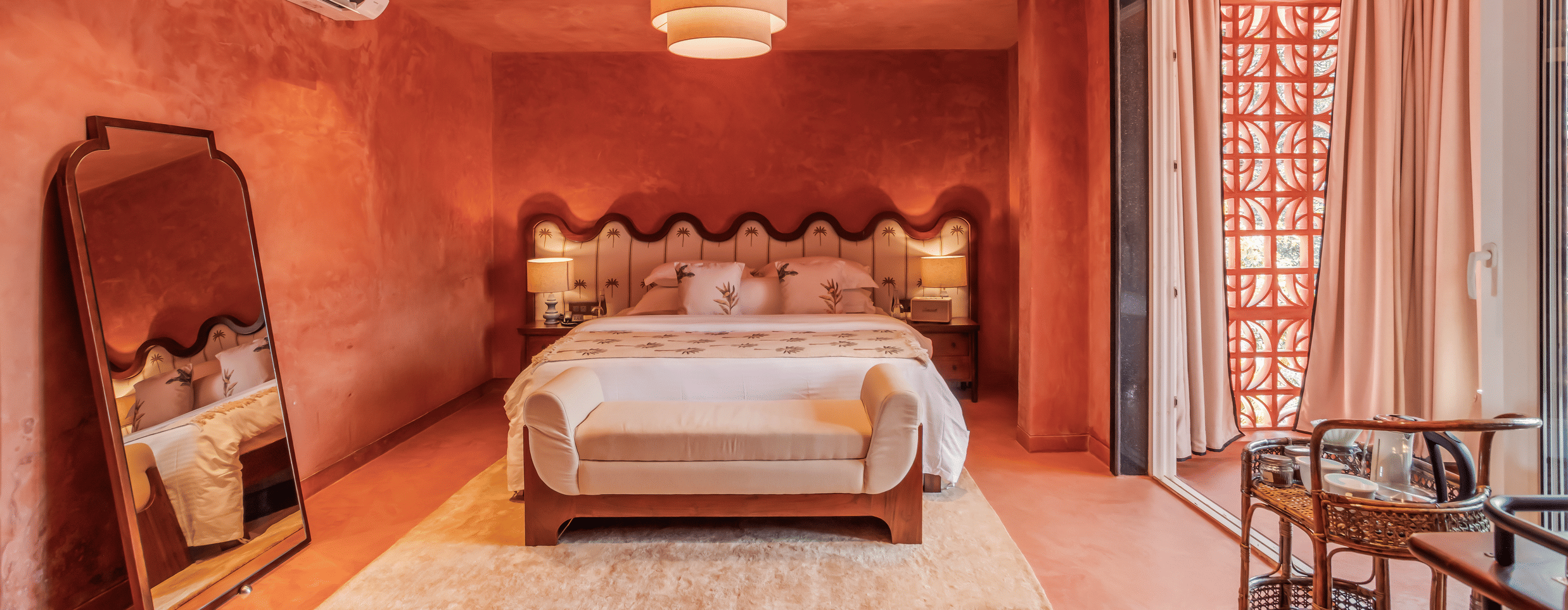 Bedroom at Unseen Boutique Hotel with reddish?orange walls, scalloped headboard, white bedding, pendant ceiling light, large mirror, and sliding door to balcony.