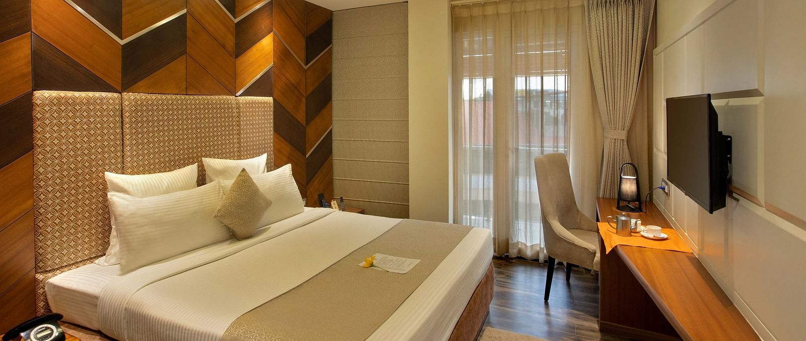 Image of warmly lit Coral Suite at Coral Reef Hotel and Spa in Port Blair featuring a plush bed in front of a TV mounted on the wall and next to it is a work desk and a chair near the door to balcony