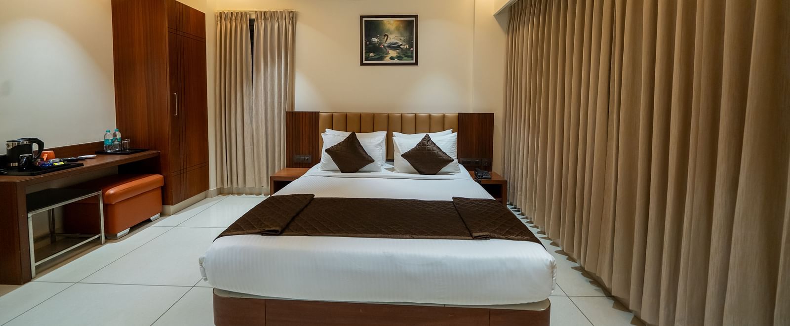 A front view of the bed at Hari Govinda's Express, Vijayanagar, with a photo frame above the headboard. To the left, a wooden cupboard stands beside a table with essentials, while on the right, an AC is mounted above the fully drawn curtains.