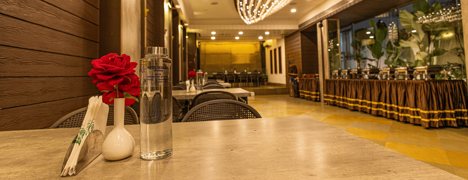 A dining area at Hotel Seagull Digha with a table, a flower in a vase, chairs, ceiling lights, and a decorative chandelier overhead.