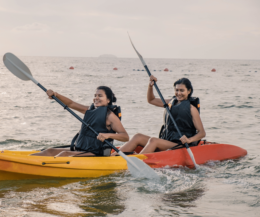 2 women in separate kayaks on the sea, paddling with smiles, enjoying a beautiful sunset, reflecting a calm and joyful water activity.