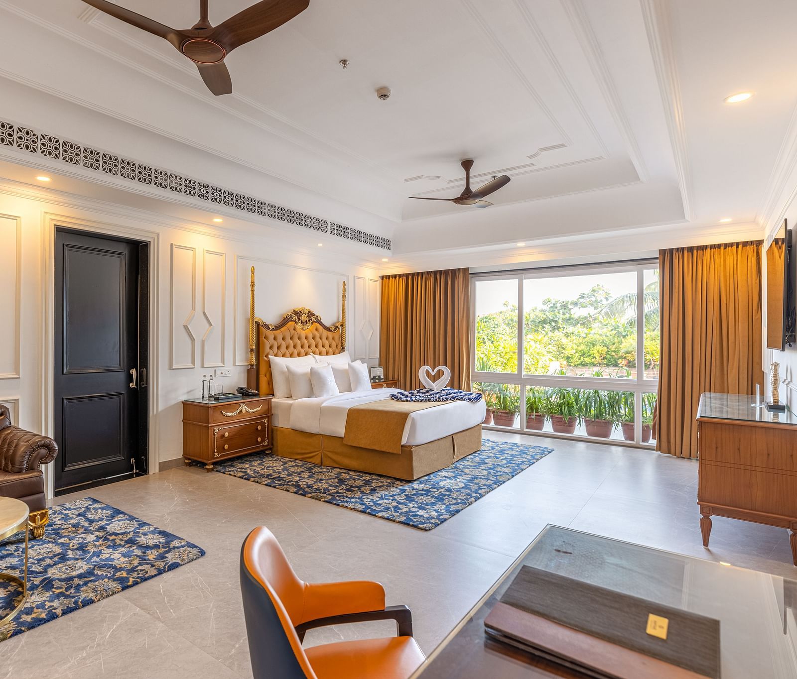 Spacious bedroom with workstation, lounge area, wall-mounted TV, bedside table, two ceiling fans, and large glass windows at MAYFAIR Palm Beach Resort, Gopalpur-On-Sea