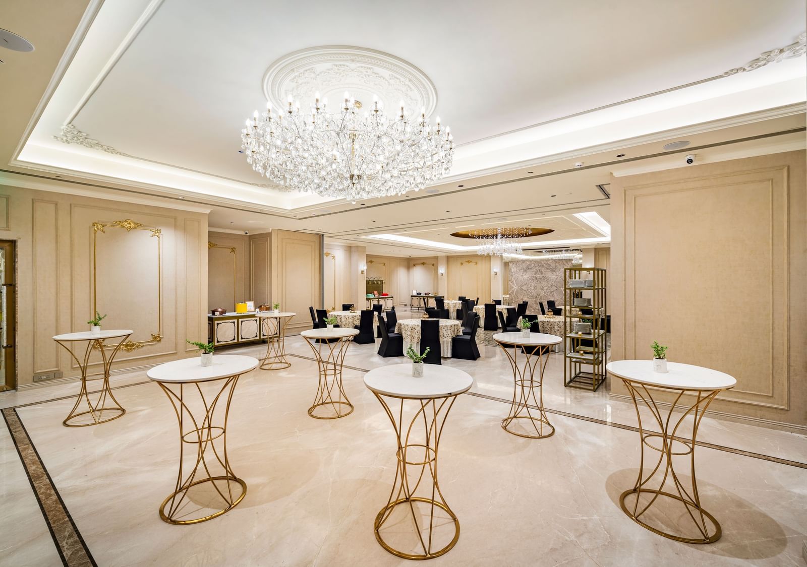 A sophisticated reception area at Grand Chennai at  several tall, ornate cocktail tables scattered across a patterned floor. A large, spherical chandelier hangs centrally, casting a soft glow over the inviting space, ideal for mingling.