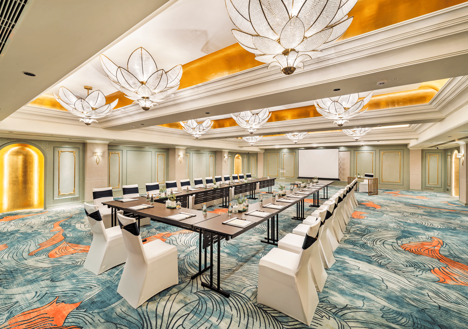 A refined meeting room at Grand Chennai by GRT Hotels centred around a substantial dark rectangular table, surrounded by modern white chairs, perfect for corporate discussions, all bathed in the light of elaborate ceiling fixtures.