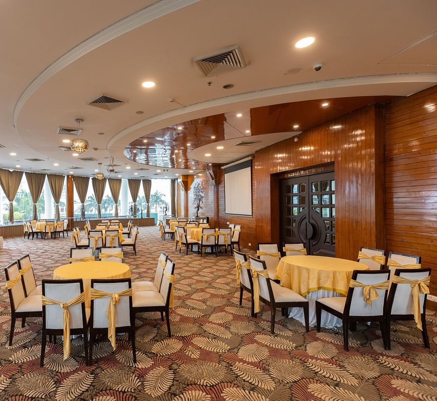Elegant indoor banquet hall at Pride Premier Dehradun with floral stage decor, round dining tables dressed in yellow linens, and large windows offering natural light—perfect for weddings or special events.