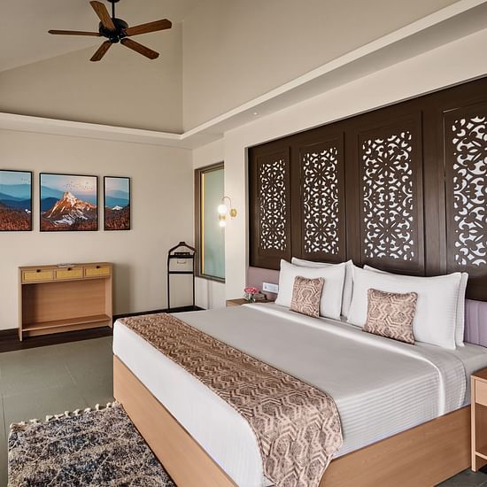 Stylish hotel bedroom with a large bed, decorative wooden panel headboard, ceiling fan, framed artwork on the wall, and floor-to-ceiling curtains - c