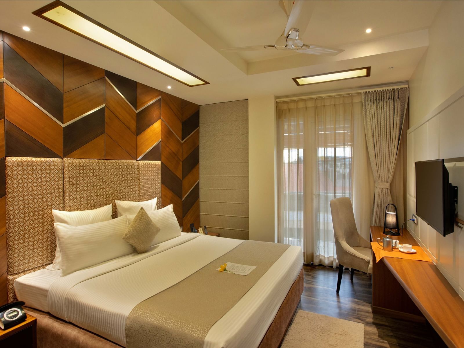 Image of warmly lit Coral Suite at Coral Reef Hotel and Spa in Port Blair featuring a plush bed in front of a TV mounted on the wall and next to it is a work desk and a chair near the door to balcony