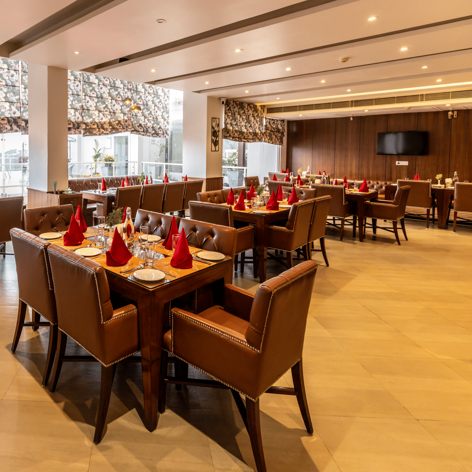 A restaurant with tables, chairs, and large windows allowing light inside at Indraprastha Resort & Spa, McLeod Ganj.