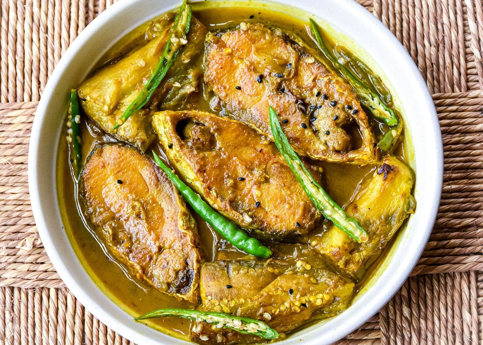 A bowl of fish curry with green chilies on a textured surface.