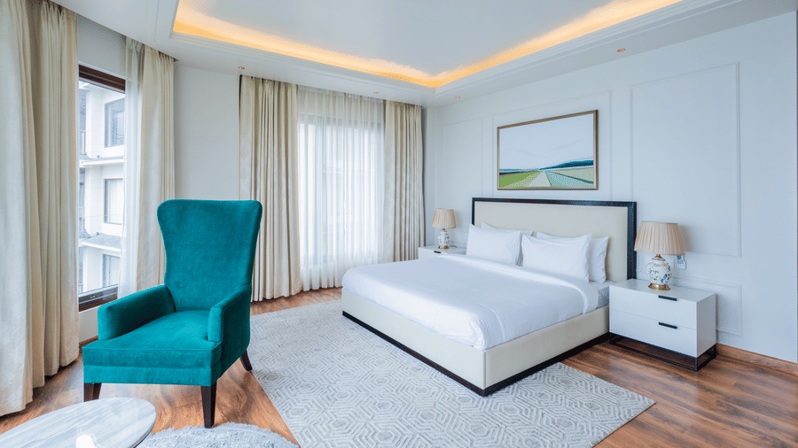 Bright, airy bedroom decorated in white tones with accent furniture and expansive windows, reflecting the stylish and contemporary interiors found at The Manor Sports & Wellness Hotel in Shimla.