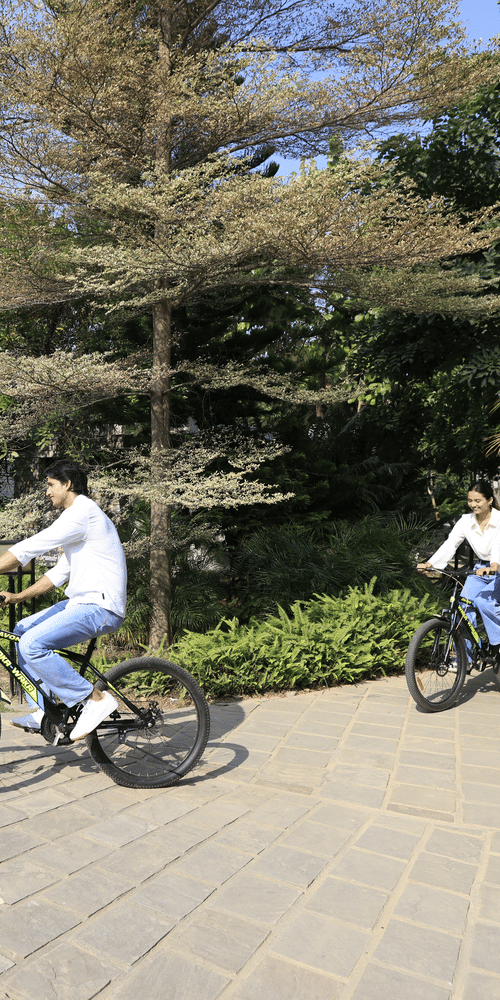 Two people riding bicycles on a paved path outdoors with trees and greenery at The Golden Tusk, Jim Corbett