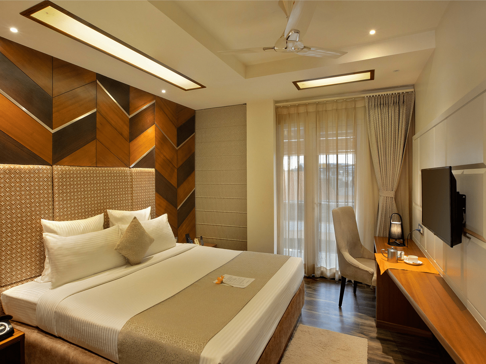 Image of warmly lit Coral Suite at Coral Reef Hotel and Spa in Port Blair featuring a plush bed in front of a TV mounted on the wall and next to it is a work desk and a chair near the door to balcony
