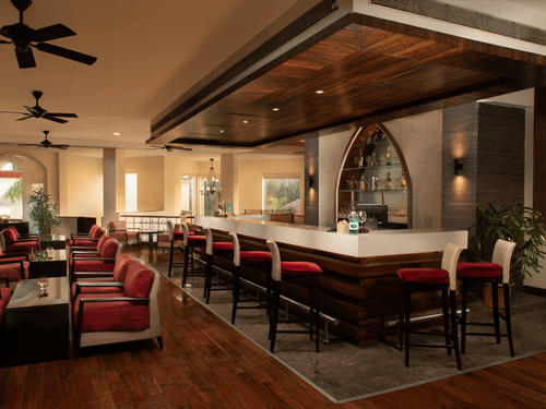 An overview of the Lobby Bar at Kenilworth Resort & Spa, Goa, with premium seating arrangements, ceiling fans, a bar counter and many drinks kept behind it.