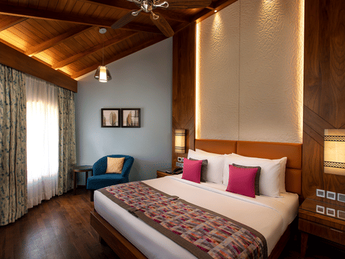 An overview of the Presidential Suite with a king bed, white and red pillows, a blue chair, paintings on the wall, accent lights above the headboard, and side tables and lamps next to the bed at Kenilworth Resort & Spa, Goa.