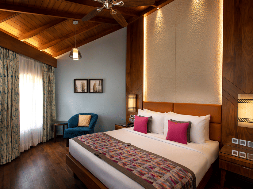 An overview of the Presidential Suite with a king bed, white and red pillows, a blue chair, paintings on the wall, accent lights above the headboard, and side tables and lamps next to the bed at Kenilworth Resort & Spa, Goa.