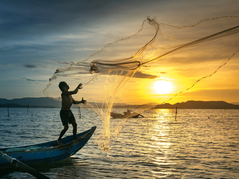 A fisherman throwing a fishing net in the sea while being on the edge of his boat with the setting sun in the background.