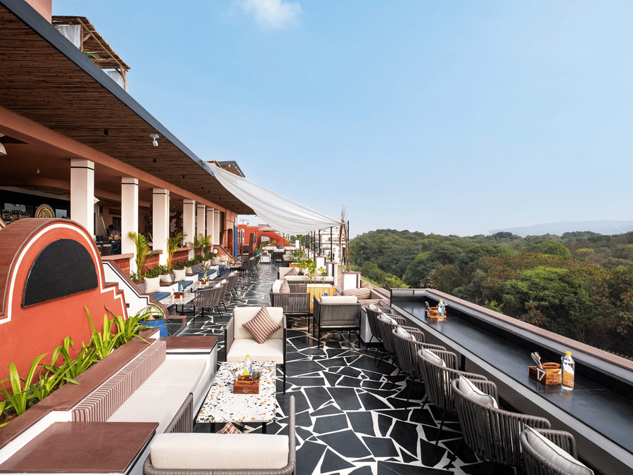 A rooftop terrace with chic outdoor furniture, a striking black and white floor, and lush greenery in the background at The Sierra, Udaipur.