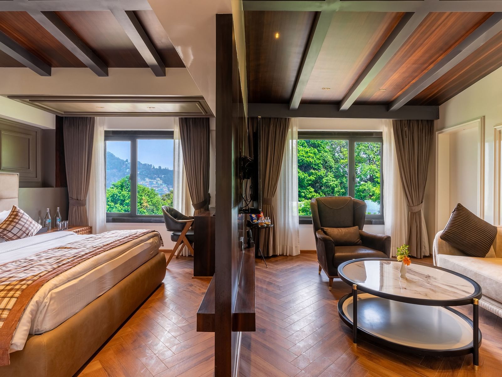 An image of a bedroom with partition, on one side is a king size bed, a chair and table and on the other side is a seating area with sofa set and a centre table