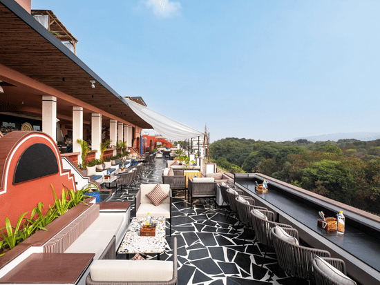 A rooftop terrace with chic outdoor furniture, a striking black and white floor, and lush greenery in the background at The Sierra, Udaipur.