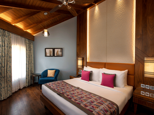 An overview of the Presidential Suite with a king bed, white and red pillows, a blue chair, paintings on the wall, accent lights above the headboard, and side tables and lamps next to the bed at Kenilworth Resort & Spa, Goa, the ideal place for a stay in G