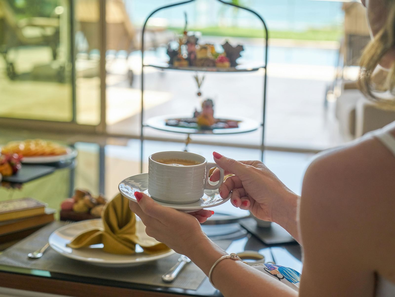 A person enjoying a high tea experience, with a tiered tray of treats and a cup of tea in hand.