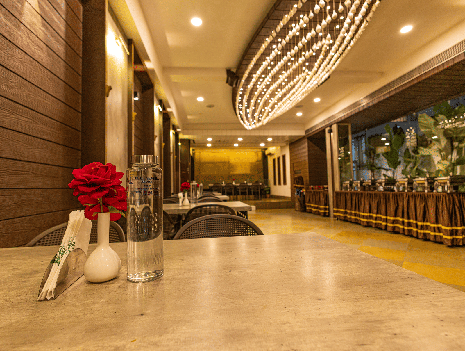 A dining area at Hotel Seagull Digha with a table, a flower in a vase, chairs, ceiling lights, and a decorative chandelier overhead.