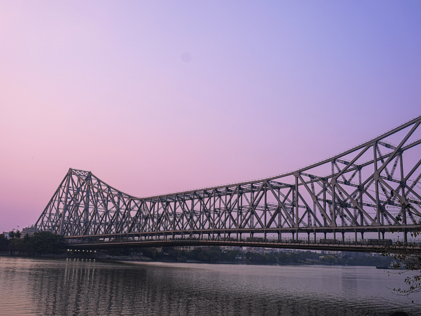 Howrah Bridge at dusk against a soft pink sky, its silhouette stretching across the river in a calm, reflective setting