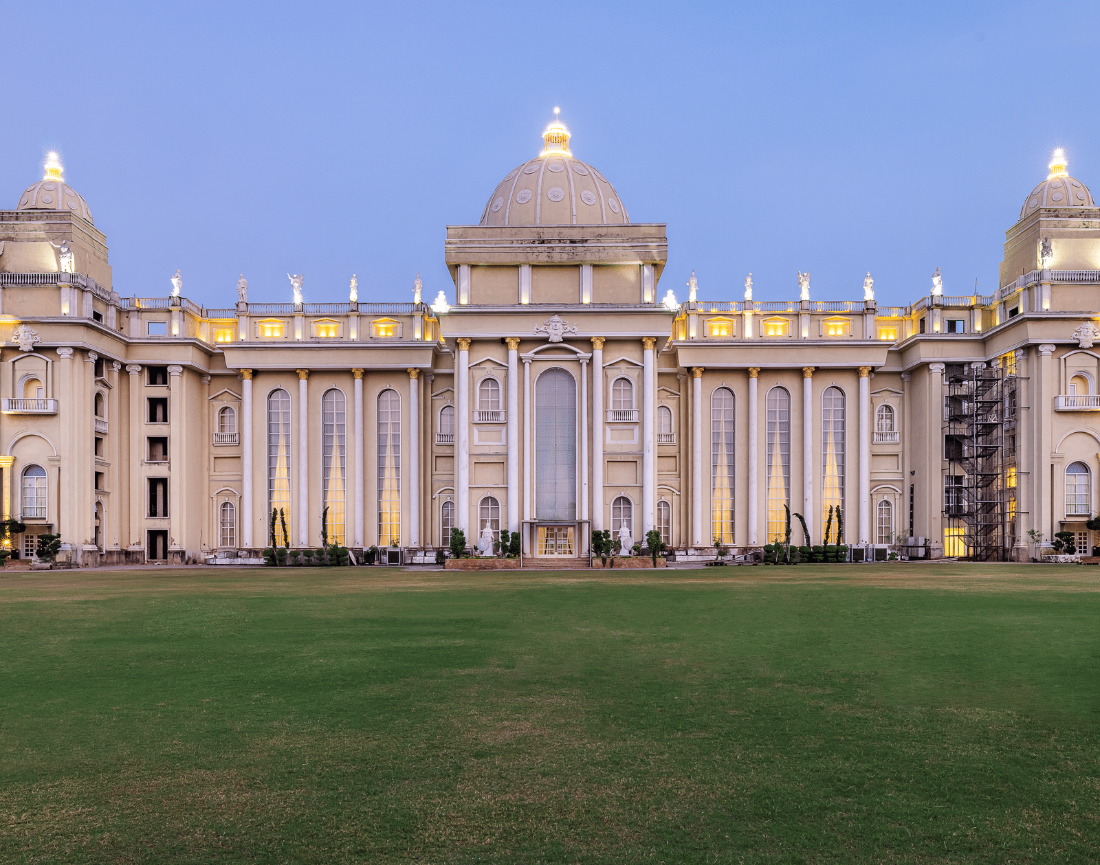 Vast, ornate, pale-coloured building with a central dome and grand architecture, illuminated against a twilight sky, set on a lawn at Hotel Hukam's Lalit Mahal.