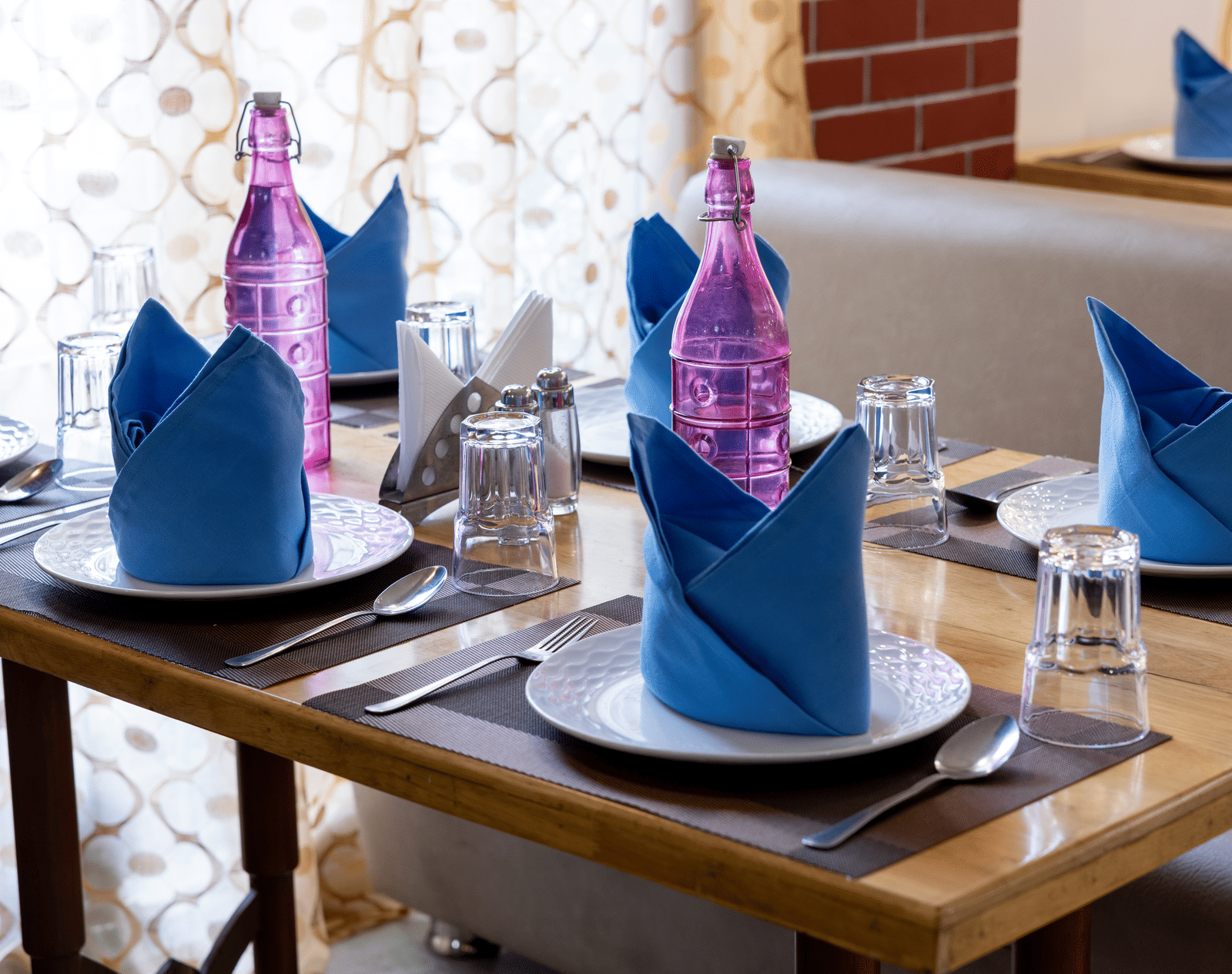 Close-up of a wooden table setting with white plates and striking blue and pink folded napkins in a well-lit dining area at Pravasa Stay, Vadodara.