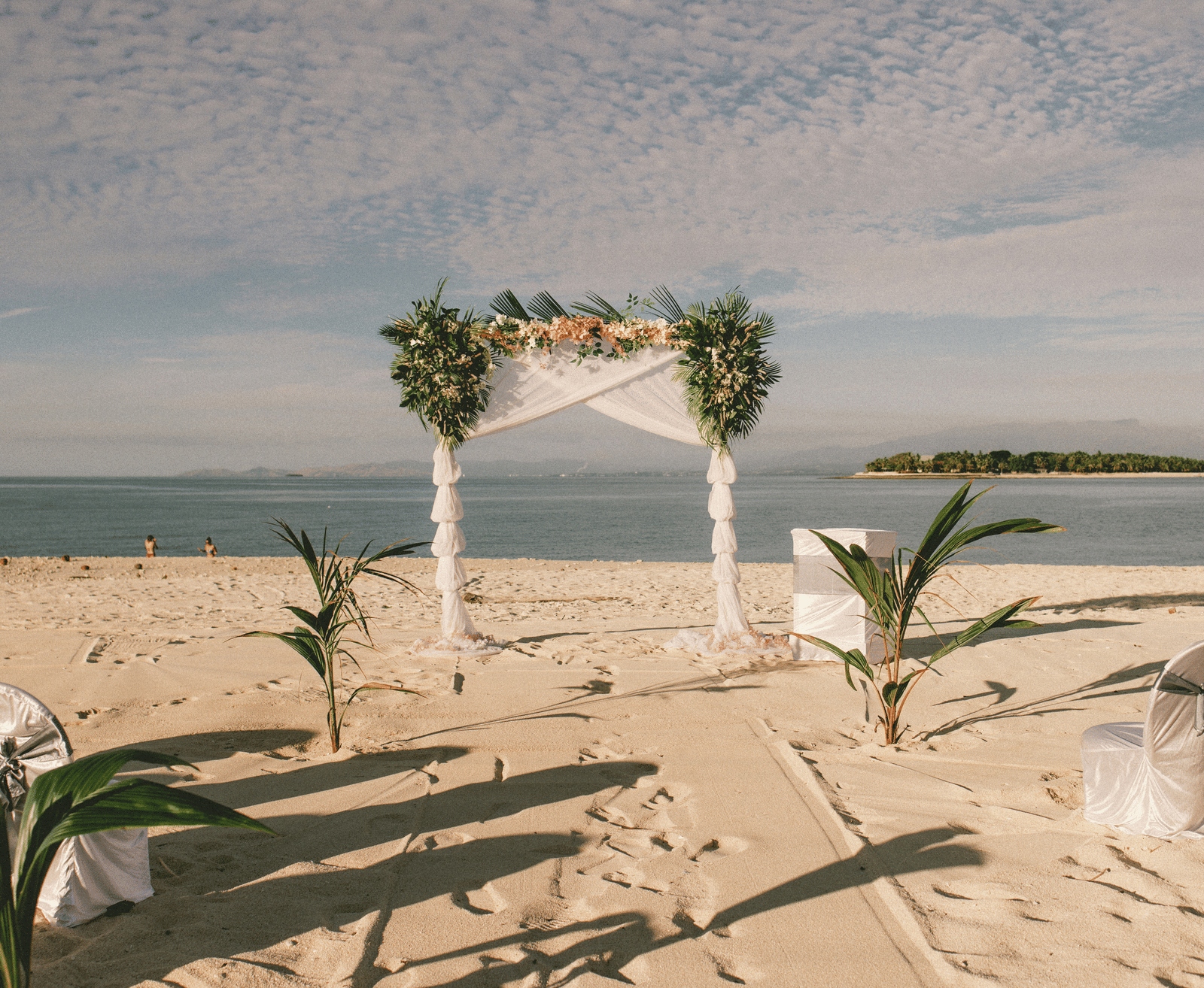 A tropical beach wedding setup with a white canopy arch decorated with foliage and facing the sea.