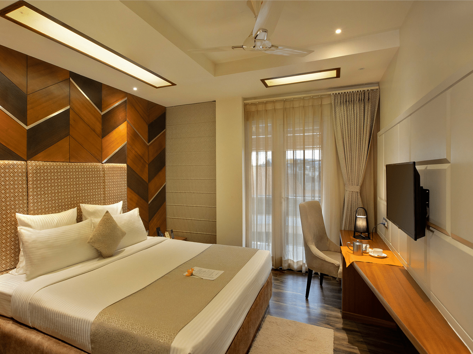 Image of warmly lit Coral Suite at Coral Reef Hotel and Spa in Port Blair featuring a plush bed in front of a TV mounted on the wall and next to it is a work desk and a chair near the door to balcony