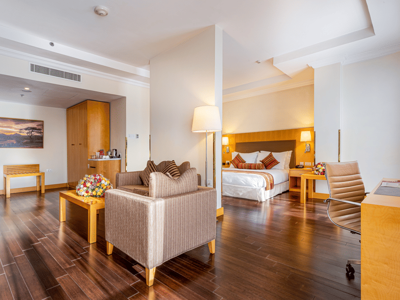 A suite with a bed, seating area, desk, chairs, tables, lamps, wooden floor, wall art, wardrobe, and a tea and coffee setup at Ramada by Wyndham Addis, Addis Ababa.
