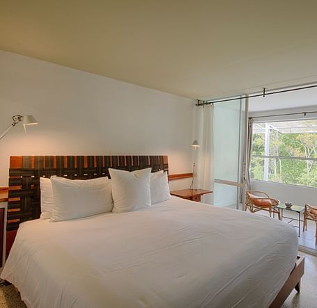 Bed with white linens at Casa Morada, facing a window with a garden view and two chairs.