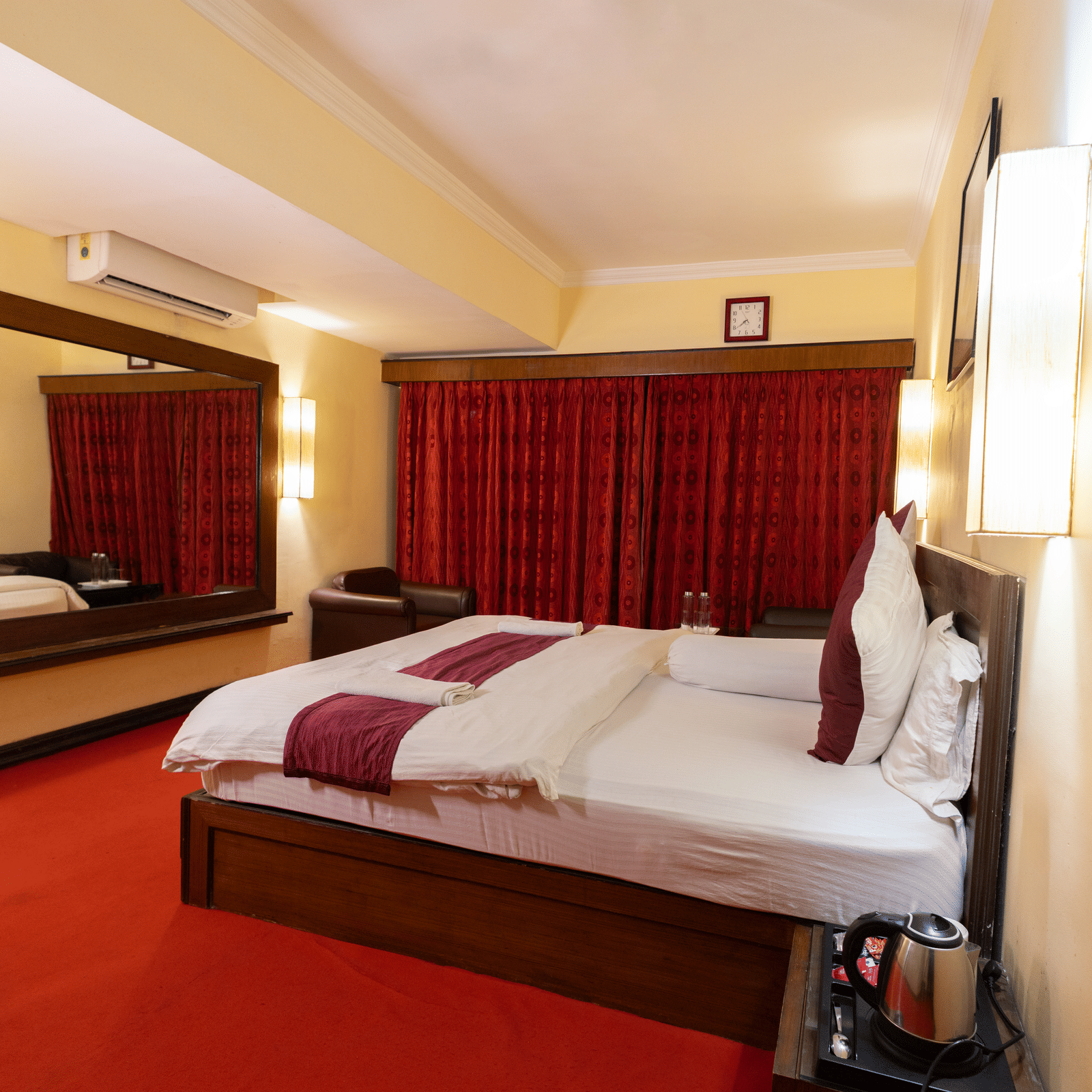 A bedroom with a king size bed, an armchair, a mirror, an A/C, and red carpeting at Hotel Seagull Digha.