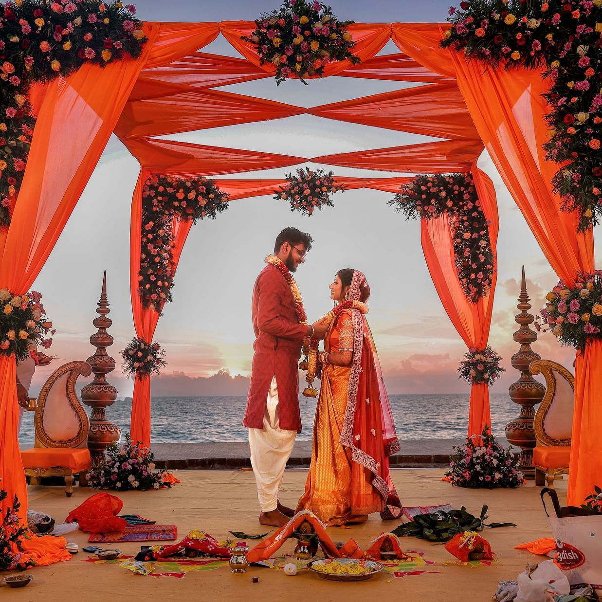 An image of a couple standing together in the wedding mandap by the beach at sunset, with a beautifully decorated floral design as they exchange vows
