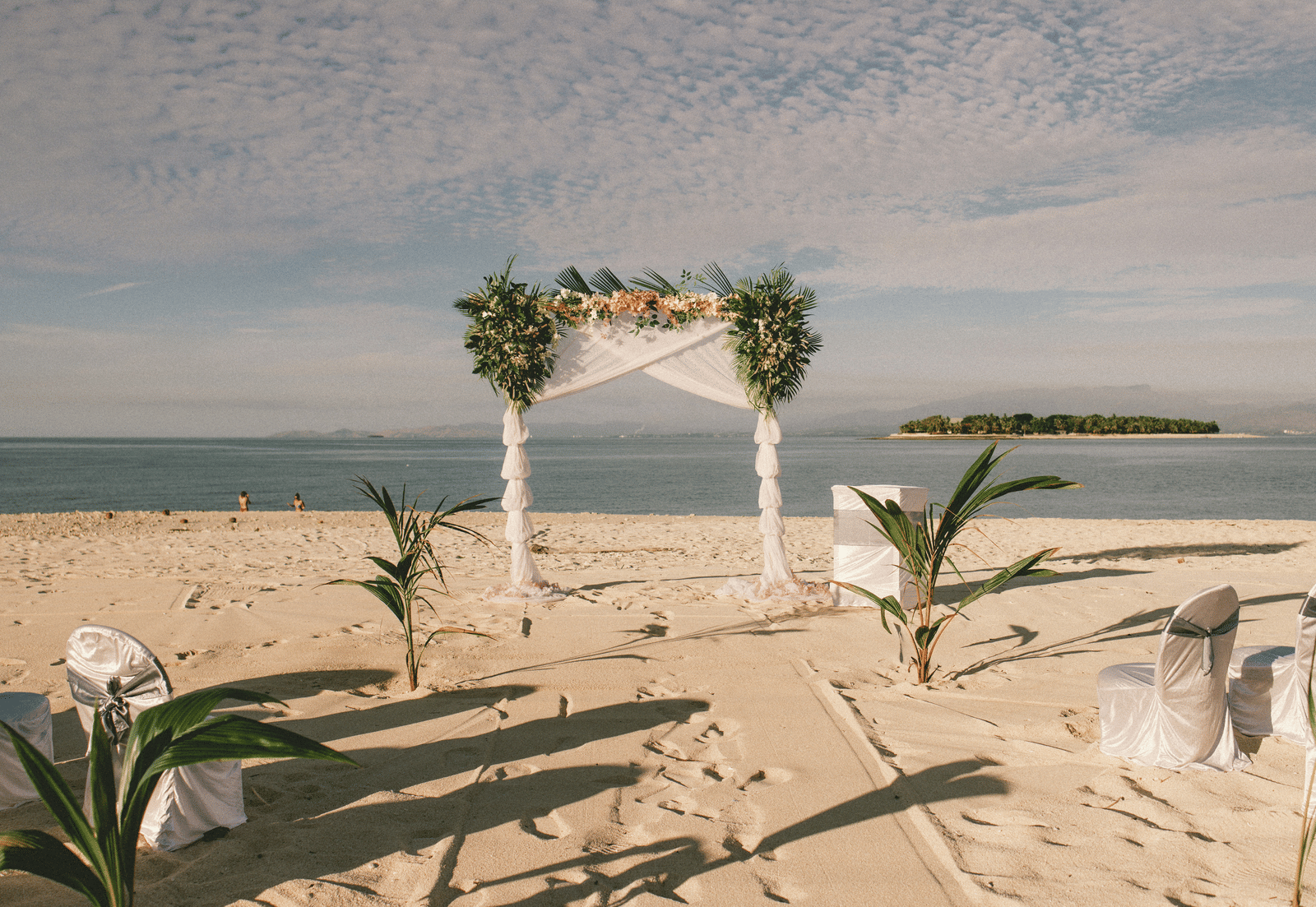 A tropical beach wedding setup with a white canopy arch decorated with foliage and facing the sea.