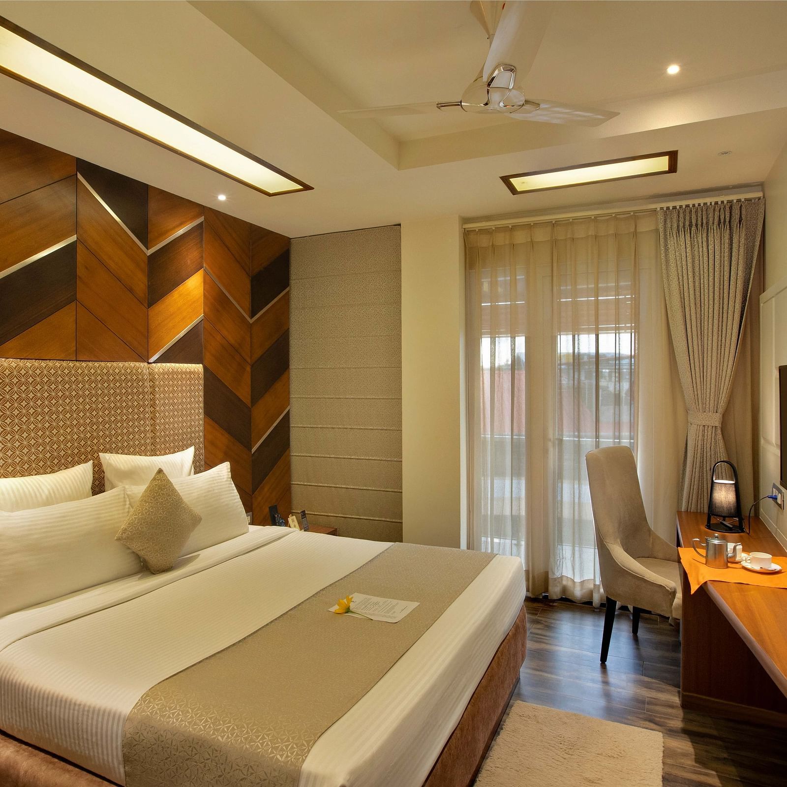 Image of warmly lit Coral Suite at Coral Reef Hotel and Spa in Port Blair featuring a plush bed in front of a TV mounted on the wall and next to it is a work desk and a chair near the door to balcony