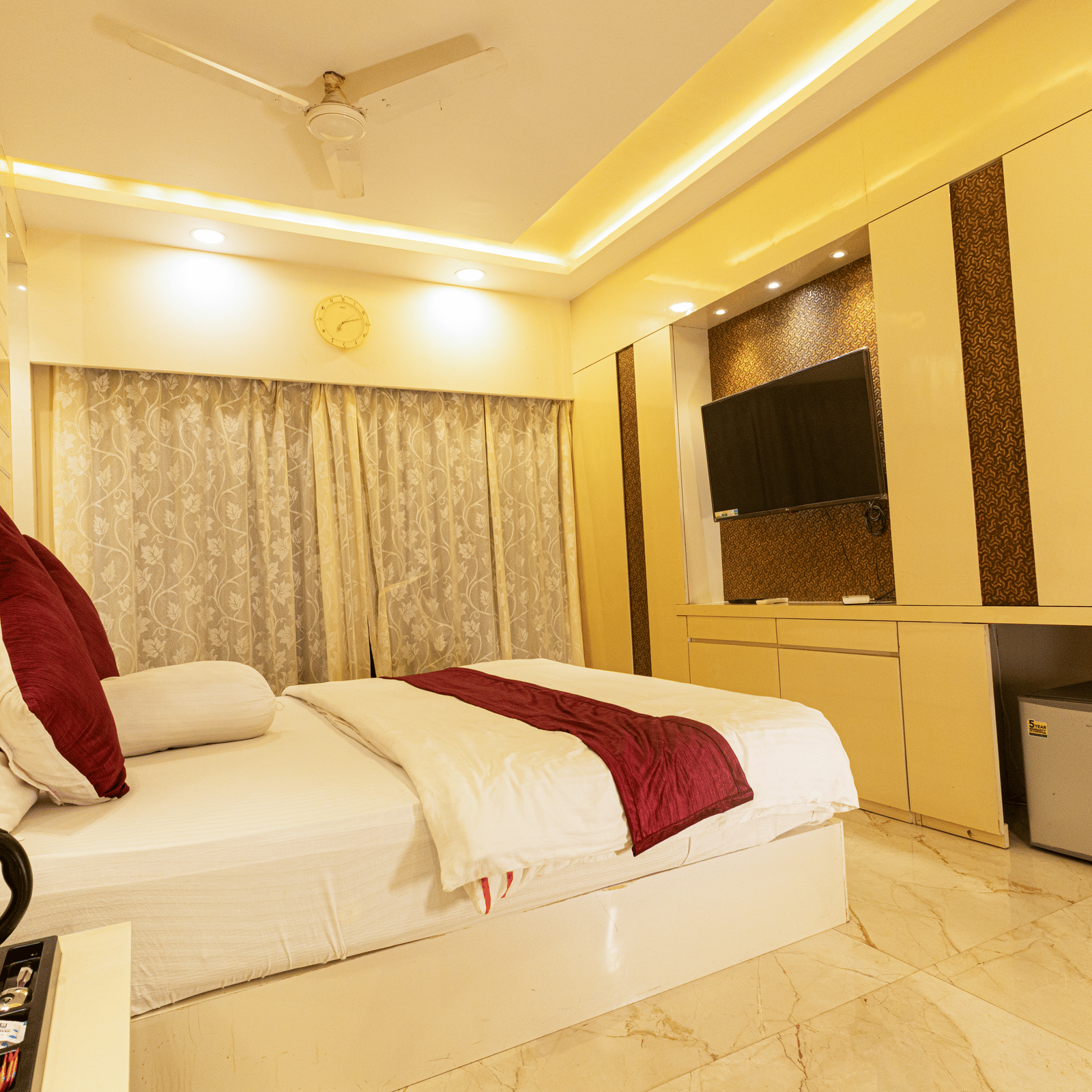 A bedroom with a king size bed, a bedside table with an electric kettle, a television, a mini fridge, and curtains at Hotel Seagull Digha.