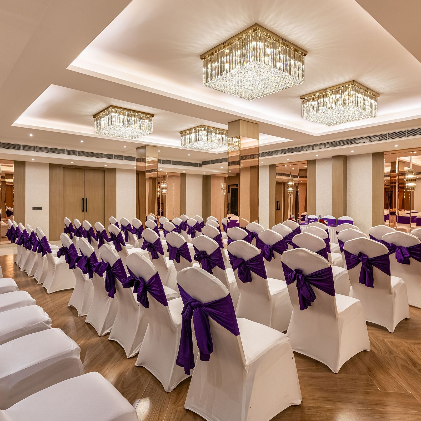 An event space with rows of white chairs adorned with purple fabric, ready for guests. It features a decorative ceiling with hanging lights and mirrored wall accents - Lords Eco Inn Dehradun.