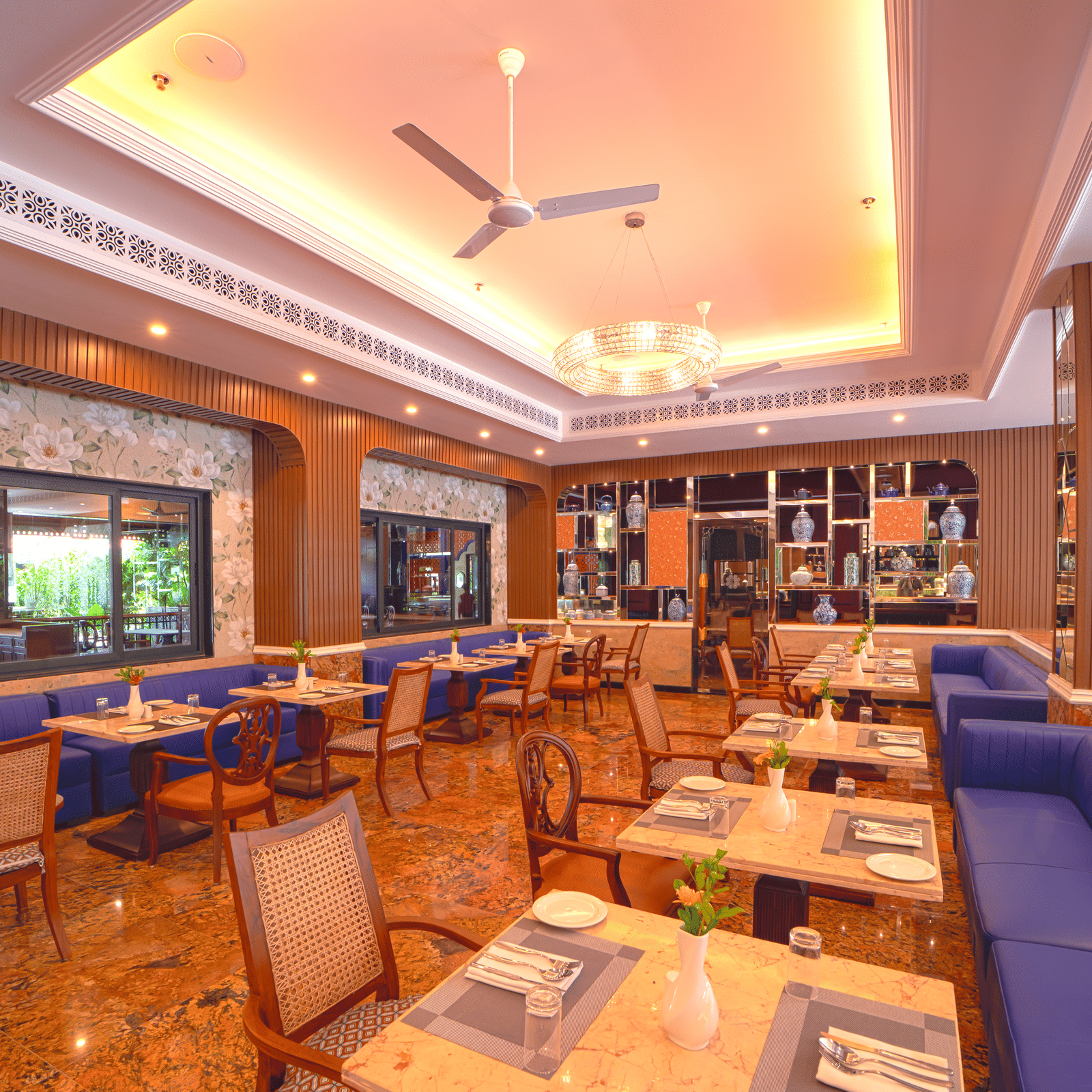 A modern dining space featuring blue cushioned seating, neatly arranged tables, ceiling fans, and large windows offering outdoor views at MAYFAIR Bay Resort, Paradeep.