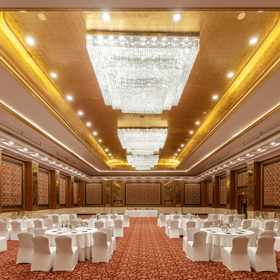 Ballroom featuring a large banquet hall at MAYFAIR Palm Beach Resort, Gopalpur-on-Sea. Chandeliers hang from the patterned ceiling. Rows of dining tables and chairs are arranged on the floor facing a stage area.