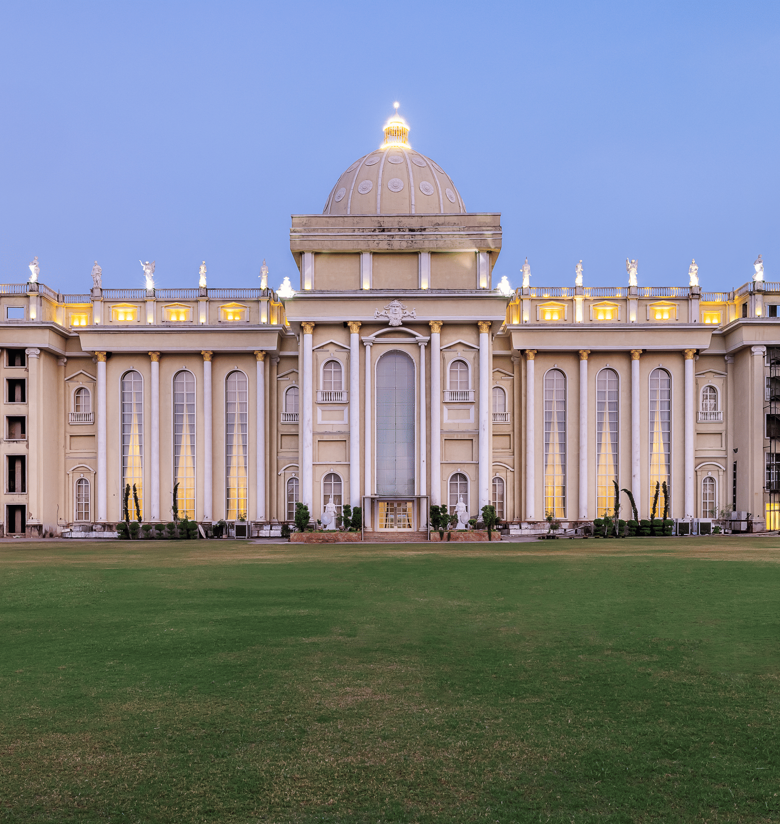 Vast, ornate, pale-coloured building with a central dome and grand architecture, illuminated against a twilight sky, set on a lawn at Hotel Hukam's Lalit Mahal.