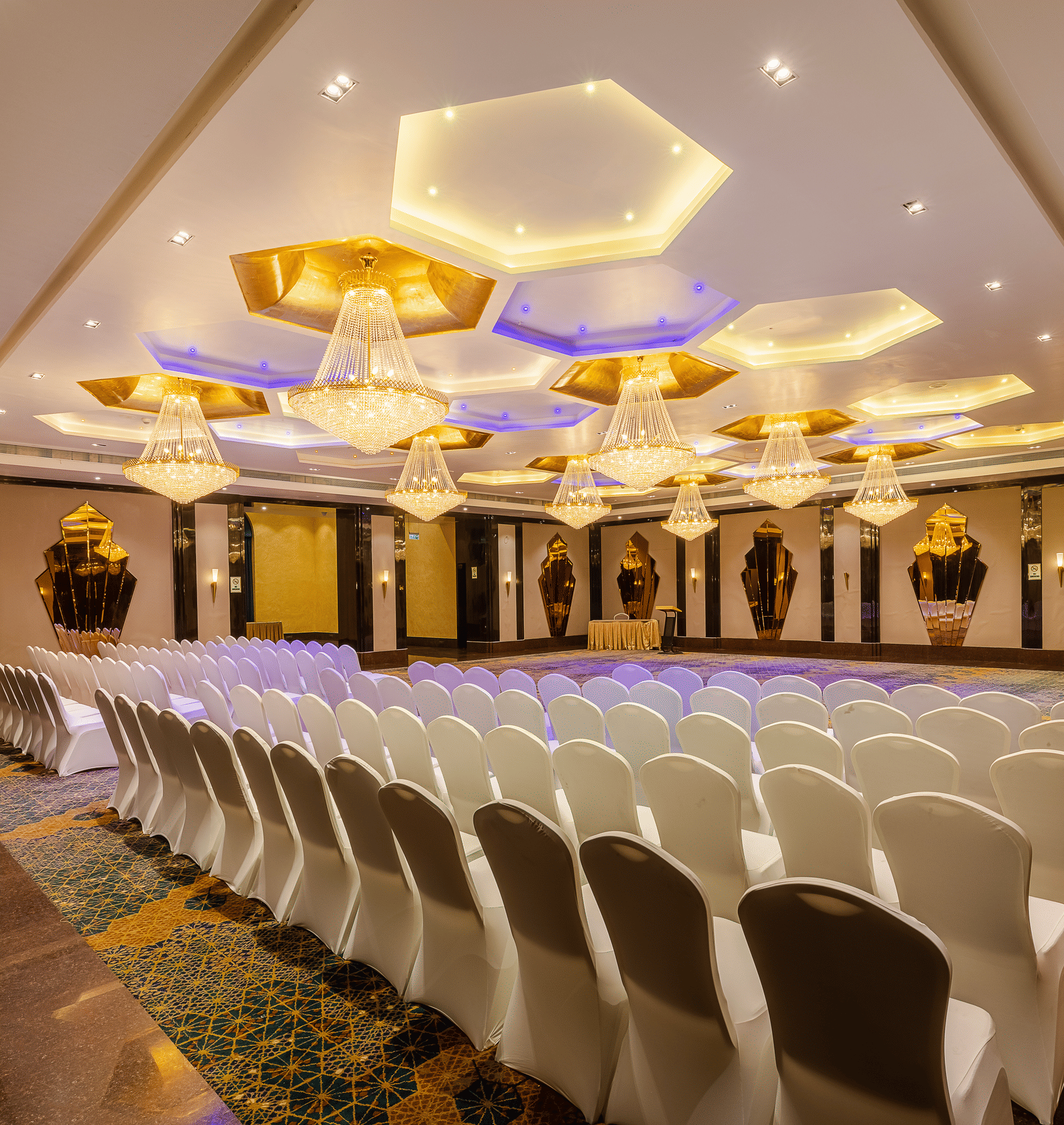 Banquet hall set with rows of white chairs facing a stage area, brightly lit by impressive hexagonal ceiling fixtures at Hotel Hukam's Lalit Mahal.