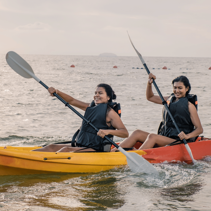 2 women in separate kayaks on the sea, paddling with smiles, enjoying a beautiful sunset, reflecting a calm and joyful water activity.