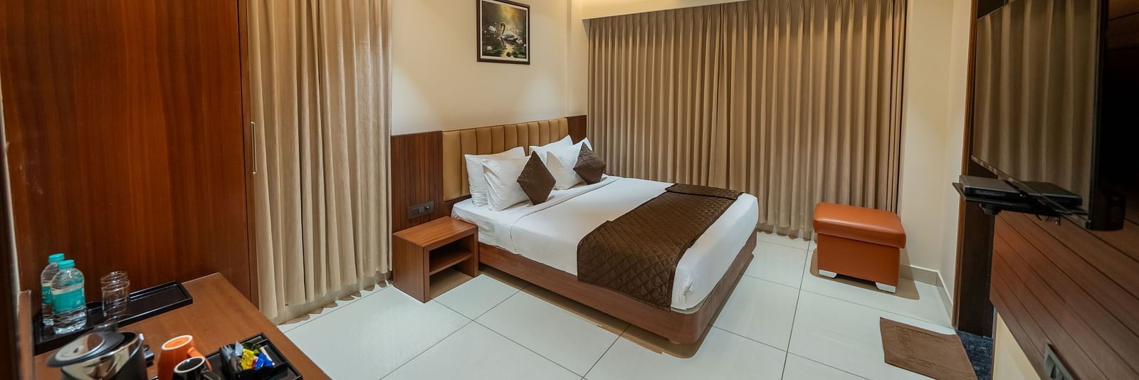 A spacious room at Hari Govinda's Express, Vijayanagar, featuring a large bed with a wooden headboard, a bedside table, a wall-mounted TV, and a wooden table with essentials, with curtains fully drawn to the side.